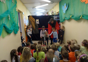 Na scenie stoi pochylony mężczyzna w stroju iluzjonisty wraz z nim stoi siedmioro dzieci. Plecami opartymi o ścianę stoi dziewczynka w granatowej sukience obejmująca chłopca. Obok iluzjonisty stoi dziewczynka w granatowej sukience. Iluzjonista ustawia dzieci (dwóch chłopców i dziewczynkę). Dziewczynka i chłopiec stoją pochyleni. Reszta dzieci siedzi na podłodze twarzami w stronę sceny.