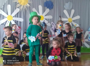 Dzieci stoją i siedzą na scenie z instrumentami. Kilkoro dzieci przebranych za pszczoły, chłopiec na pierwszym planie jest świerszczem obok niego siedzi dziewczynka biedronka.