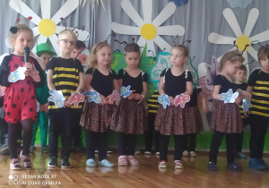 Dzieci stoją na scenie w szeregu. Dziewczynka po lewej stronie w stroju biedronki trzyma w dłoniach mikrofon. Obok niej stoi chłopiec w stroju pszczoły oraz dziewczynki motylki. Każde z dzieci ma na ręku bransoletkę z motylkiem