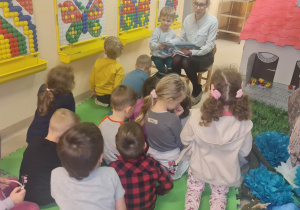 Kobieta w białej koszuli i okularach siedzi na niskim krzesełku, twarzą w stronę dzieci w rękach trzyma książkę. Po jej prawej stronie klęczy chłopiec. Pozostałe dzieci na zdjęciu widoczne plecami z twarzami w stronę pani. Zdjęcie ukazuje hol przedszkola.