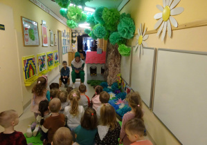 W holu przedszkola widać zielone drzewo i domek - dekoracja. Na krzesełku twarzą do dzieci siedzi kobieta w zielonym dresie i zielonej czapce na głowie. Obok niej na krześle siedzi chłopiec. Kobieta i dziecko siedzą twarzami w stronę dzieci siedzących na podłodze. Kobieta trzyma w ręku książkę.