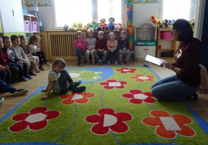 Dzieci siedzą na ławeczkach. Jeden chłopiec siedzi na zielonym dywanie w czerwone kwiaty. Wszystkie dzieci mają twarze skierowane w kierunku osoby robiącej zdjęcie. Kobieta klęka na dywanie trzymając w dłoniach otwartą książkę.