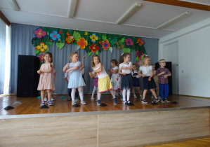 Dzieci stoją na scenie w dwóch szeregach. Dzieci w pierwszym szeregu wykonują ruch "młynka".