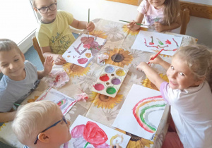 Pięcioro dzieci siedzi przy stole. Uśmiechająca się dziewczynka w białej bluzce ma namalowaną tęczę. Chłopiec w okularach maluje czerwoną farbą.