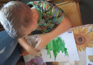 Chłopczyk w zielonej bluzce siedzi przy stole i maluje na kartonie zieloną farbą.