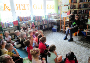 Dzieci siedzą na dywanie w bibliotece miejskiej, podnoszą ręce zgłaszając się do odpowiedzi