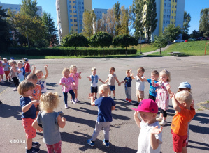 Dzieci na powietrzu stoją w kole. Mają podniesione ręce naśladują ruchem słowa piosenki