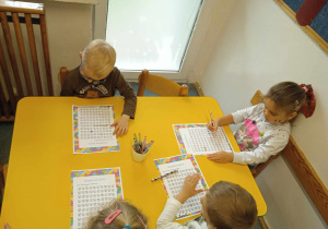 Dzieci siedzą przy żółtym stoliku i wykonują kartę pracy