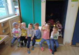 Dzieci siedzą na ławce i trzymają uśmiechnięte buźki na patyczkach