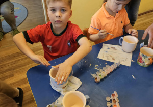 Na zdjęciu widać dzieci które malują kubki farbami na warsztatach ceramicznych.
