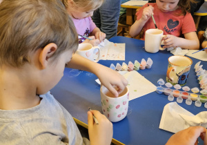 Na zdjęciu widać dzieci które malują kubki farbami na warsztatach ceramicznych.