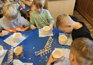 Na zdjęciu widać dzieci które malują kubki farbami na warsztatach ceramicznych.
