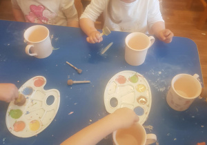 Na zdjęciu widać dzieci które malują kubki farbami na warsztatach ceramicznych.