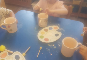 Na zdjęciu widać dzieci które malują kubki farbami na warsztatach ceramicznych.