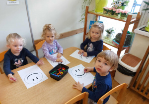 Trzy dziewczynki i chłopiec siedzą przy stoliku z twarzami zwróconymi w stronę osoby robiącej zdjęcie. Kolorują uśmiechniętą buzię.