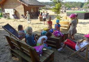 Dzieci przy stoliku na plaży wypakowują prowiant z małych plecaczków. Dwie dziewczynki siedzą na leżakach.