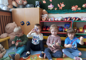 Czworo dzieci siedzi na dywanie i nawleka na druciki kreatywne koraliki. Chłopiec w białej bluzce robi z drucika koło i przykłada je do oczu.