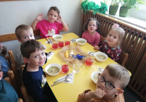 Sześcioro dzieci siedzi przy stoliku. Jedna dziewczynka dotyka rękoma do uszu. W miskach dzieci mają podaną zupę.