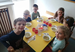 Trzy dziewczynki i trzech chłopców siedzą przy stole. Jeden chłopiec uśmiecha się pokazując zęby. Na stole stoją białe miski w których znajduje się zupa dla dzieci.