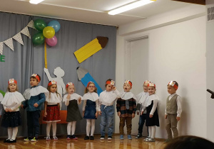 Dziesiątka dzieci w odświętnych strojach i opaskach na głowie stoi na scenie. Dzieci śpiewają lub recytują.
