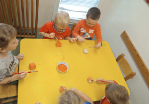Dzieci siedzą przy stoliku i malują pomarańczową farbą dynie wykonane z masy solnej.