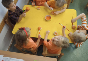 Sześcioro dzieci siedzi przy stoliku i maluje pomarańczową farbą dynie wykonane z masy solnej.