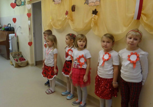 Sześcioro dzieci stoi pod ścianą w holu przedszkola. Dzieci ubrane są w barwy narodowe. Wszystkie śpiewają piosenkę.