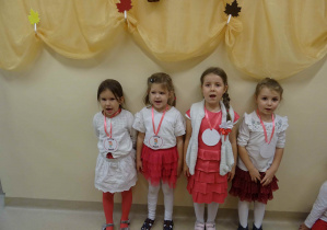 Cztery dziewczynki w barwach narodowych stoją przy ścianie. Dziewczynki mają otwarte buzie do śpiewu.