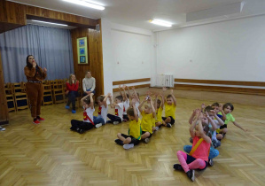 Dzieci siedzą w trzech rzędach trzymają w górze ręce