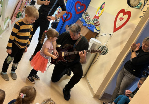 Na scenie jeden mężczyzna trzyma mikrofon. Pan z gitarą elektryczną klęczy by dziewczynka mogła dotknąć strun, dziewczynce przygląda się chłopiec stojący obok niej.