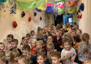 Duża grupa dzieci tańcząca na holu przedszkola przyozdobionym karnawałowo.