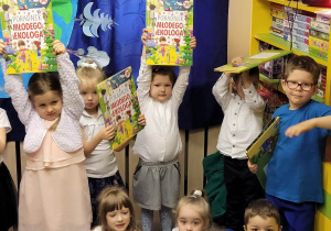 Ośmioro dzieci na zdjęciu trzyma książki nagrody, troje dzieci siedzi na podłodze i trzyma książki na kolanach, za nimi stoi piątka dzieci które trzymają książki nad głową