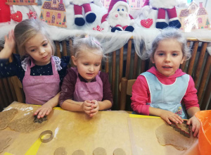 Trzy dziewczynki w fartuszkach siedzą przy stoliku i wyciskają z ciasta kształty pierników.