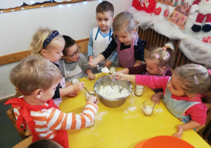 Dzieci z dużej srebrnej misy łyżeczkami przesypują mąkę do szklaneczek.