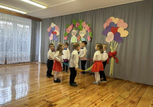 Dzieci stoją na scenie w kole trzymają w ręku związane ze sobą kolorowe szarfy W tle dekoracja z okazji dnia Babci i Dziadka oraz balony z papieru.