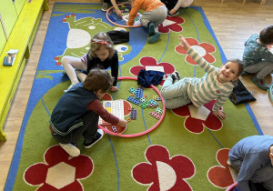 Dzieci siedzą na dywanie w grupkach trzyosobowych. W kołach hula hop znajdują się klocki Numicon