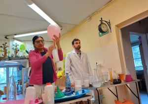 Pani w różowym swetrze i pan w białym fartuchu chemika stoją przy stole z probówkami. Kobieta trzyma w ręku balon naciągnięty na butelkę, który jest pompowany powietrzem z butelki.