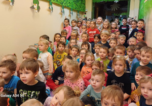 Duża grupa przedszkolaków siedzi w holu przedszkola i przygląda się pokazom chemicznym.
