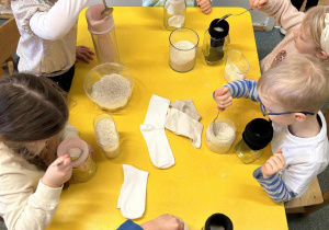Dzieci siedzą przy stolikach i wsypują ryż do skarpet