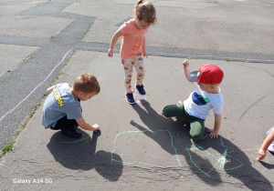 Dwóch chłopców rysuje kredą na betonowym boisku. Dziewczynka podskakuje na narysowanym przez siebie kwadracie.