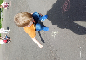 Chłopiec w blond włosach i pomarańczowej bluzeczce rysuje kredą na betonie
