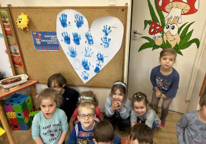 Dzieci siedzą na dywanie za nimi na tablicy wisi białe serce z odciśniętymi niebieskimi dłońmi.