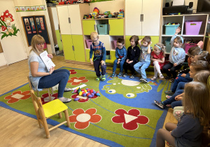 Dzieci siedzą w półkolu na ławce, słuchają opowiadania czytanego przez nauczyciela