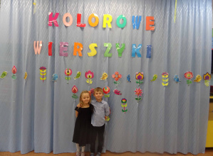 Chłopiec z dziewczynką odświętnie ubrani stoją na scenie za nimi dekoracja z napisem Kolorowe Wierszyki.
