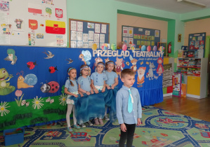 Chłopiec w niebieskiej koszuli stojąc na środku sali recytuje wiersz. Za nim stoi pięć dziewczynek ubranych na niebiesko trzymających niebieski materiał.