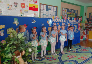 Dzieci ubrane na niebiesko stoją w szeregu i trzymają palec na ustach.